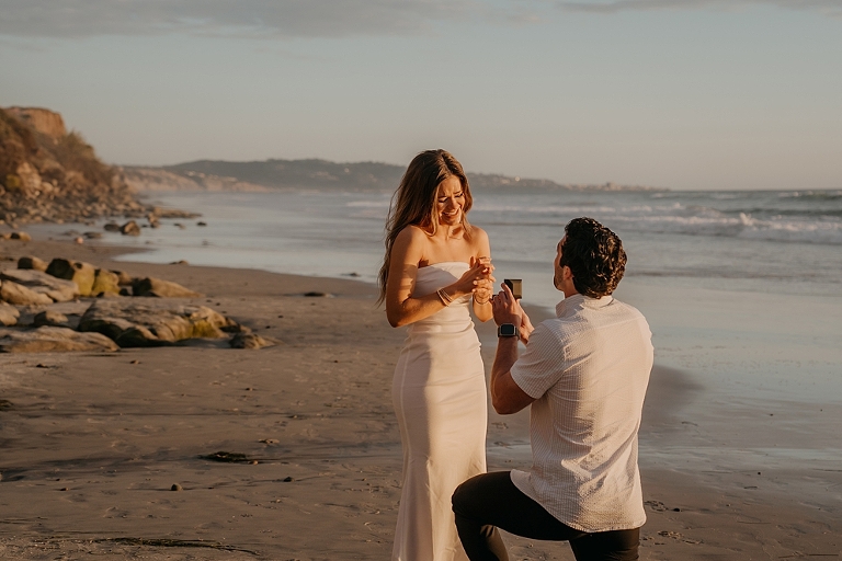 San Diego Proposal Photographer Man proposing to woman on Del Mar Beach at sunset, both smiling happily as woman extends her hand towards man.