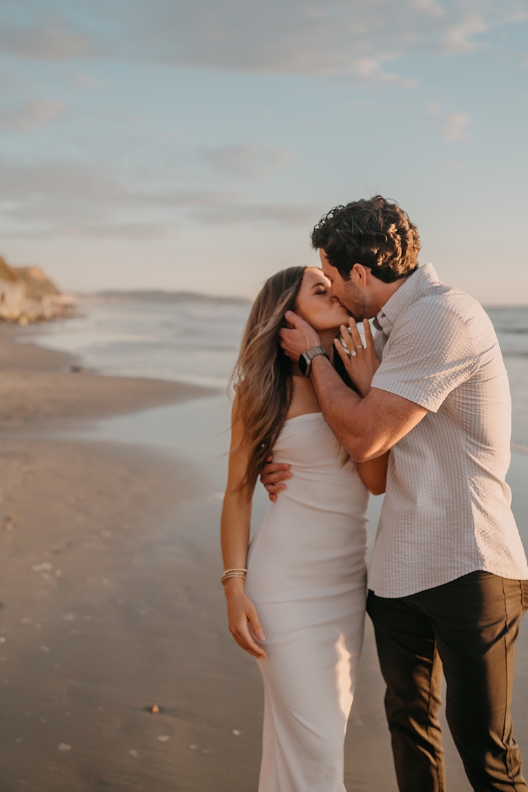 San Diego Proposal Photographer A couple kisses on Del Mar Beach at sunset, with the woman in a white dress and the man in a casual shirt and pants.