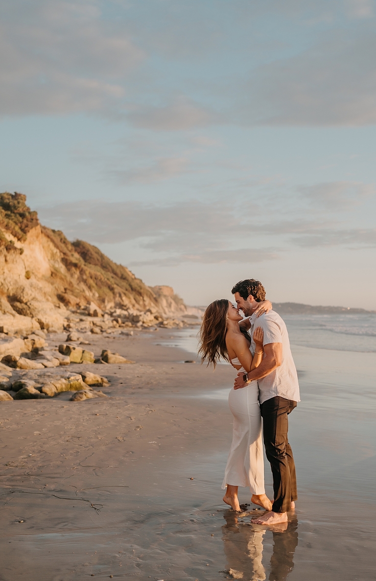 San Diego Proposal Photographer A couple kissing on Del Mar Beach at sunset, with one person in a white dress and the other in black pants and a white shirt, surrounded by rocks and cliffs.