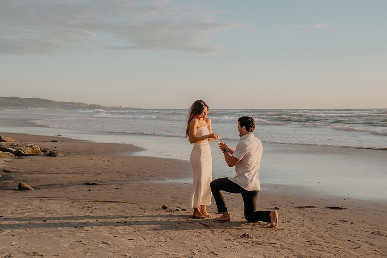 San Diego Proposal Photographer A man kneeling on Del Mar Beach proposing to a woman at sunset, with waves and cliffs in the background.