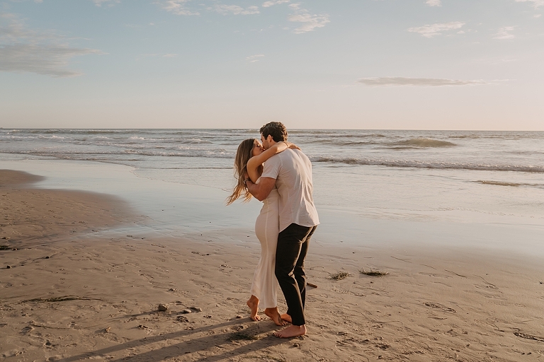 San Diego Proposal Photographer A couple embraces and kisses on the sandy Del Mar Beach at sunset, with gentle waves in the background.