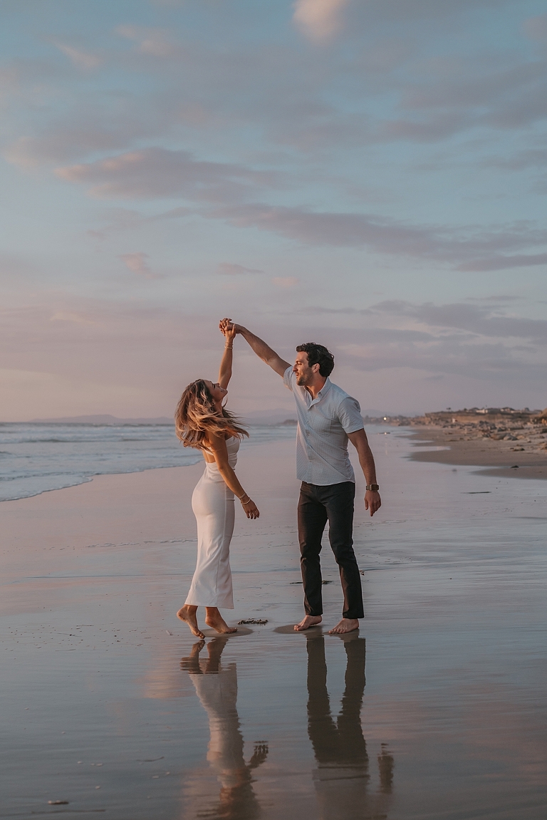 San Diego Proposal Photographer A couple joyfully dancing on a beach at sunset, the man twirling the woman by the hand, both barefoot with waves gently breaking near their feet.