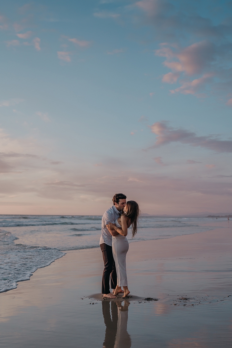 San Diego Proposal Photographer A couple embraces on Del Mar Beach at sunset, with the ocean and pink-tinted sky in the background.