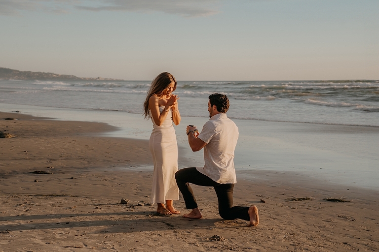 San Diego Proposal Photographer A man proposes to a woman on Del Mar Beach at sunset; she is surprised and emotional, both dressed semi-formally.