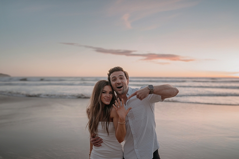 San Diego Proposal Photographer A happy couple posing with the woman embracing the man from behind on Del Mar Beach at sunset, both smiling at the camera.